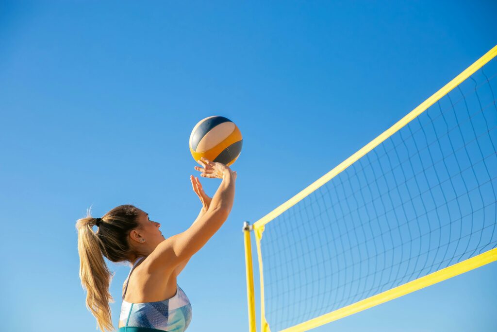 A woman plays volleyball on a sunny day, reaching to hit the ball over the net.